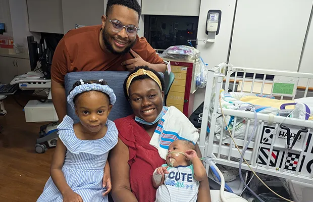 Couple holding their baby in the hospital while smiling at the camera next to a little girl
