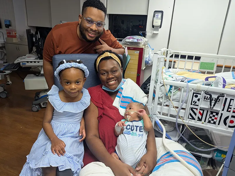 Couple holding their baby in the hospital while smiling at the camera next to a little girl