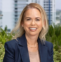 Photo of a woman smiling at the camera, wearing a dark blue suit, with a background of bushes.