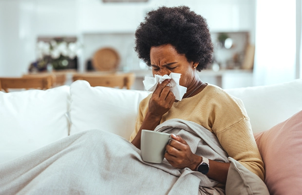 A woman sneezing while sitting on a sofa, covering her mouth with a blanket.