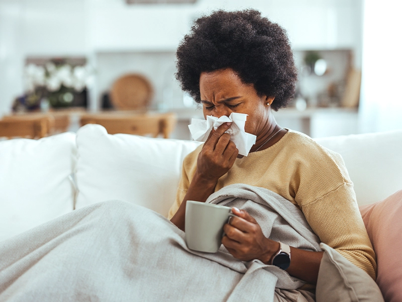 A woman sneezing while sitting on a sofa, covering her mouth with a blanket.