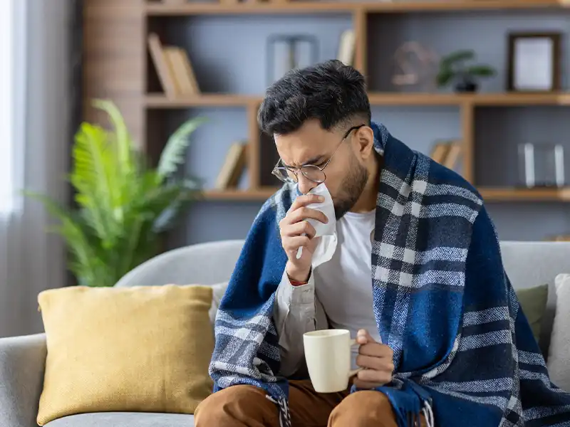 Man sitting in his living room wearing comfortable clothes and covered with a blanket while coughing