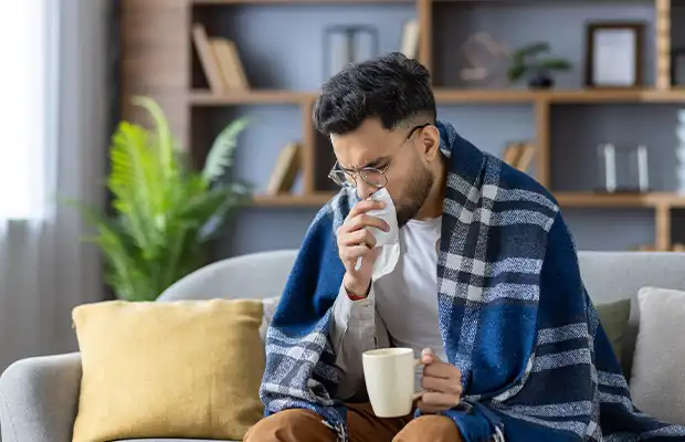 Man sitting in his living room wearing comfortable clothes and covered with a blanket while coughing