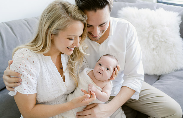 A family of three, a new mom and dad holding their baby, they are sitting on a couch