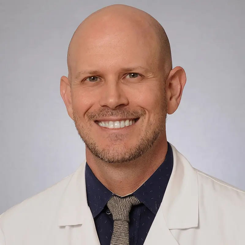 A medical professional smiling at the camera, he has on a white coat and dark blue shirt