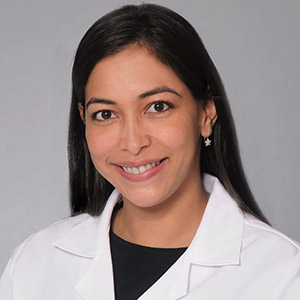 Headshot of Diana Molinares, she smiles at the camera, has long black hair, has on a black shirt and white coat