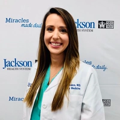 Headshot of a medical professional smiling at the camera, she has long brown hair and is wearing a white coat