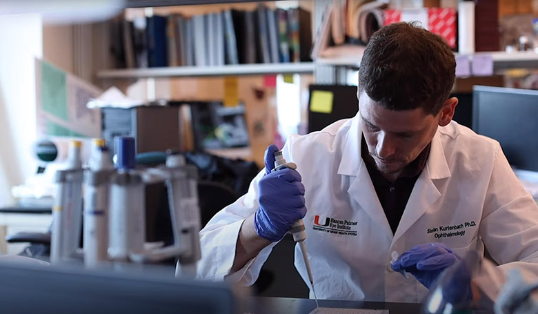Graduate student doing lab tests, a male student sits, he is wearing a lab coat and running tests