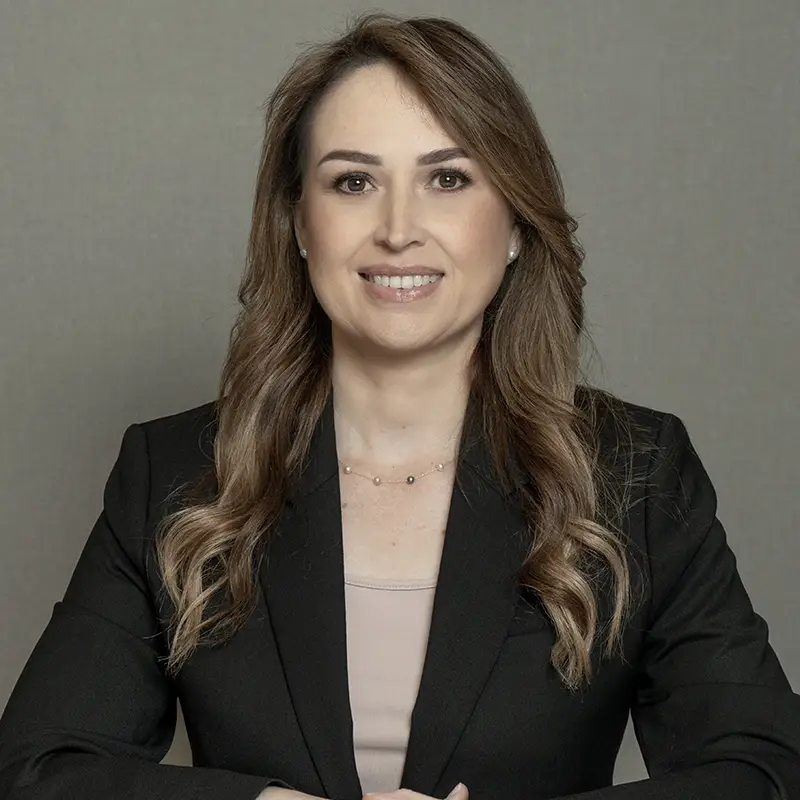 A woman smiling at the camera, she has a dark coat, brown top, and long light brown hair