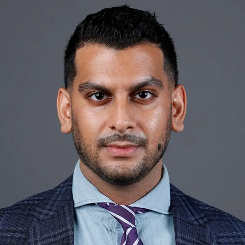 A man looking at the camera, he has a purple and white tie, a dark coat, and blue shirt