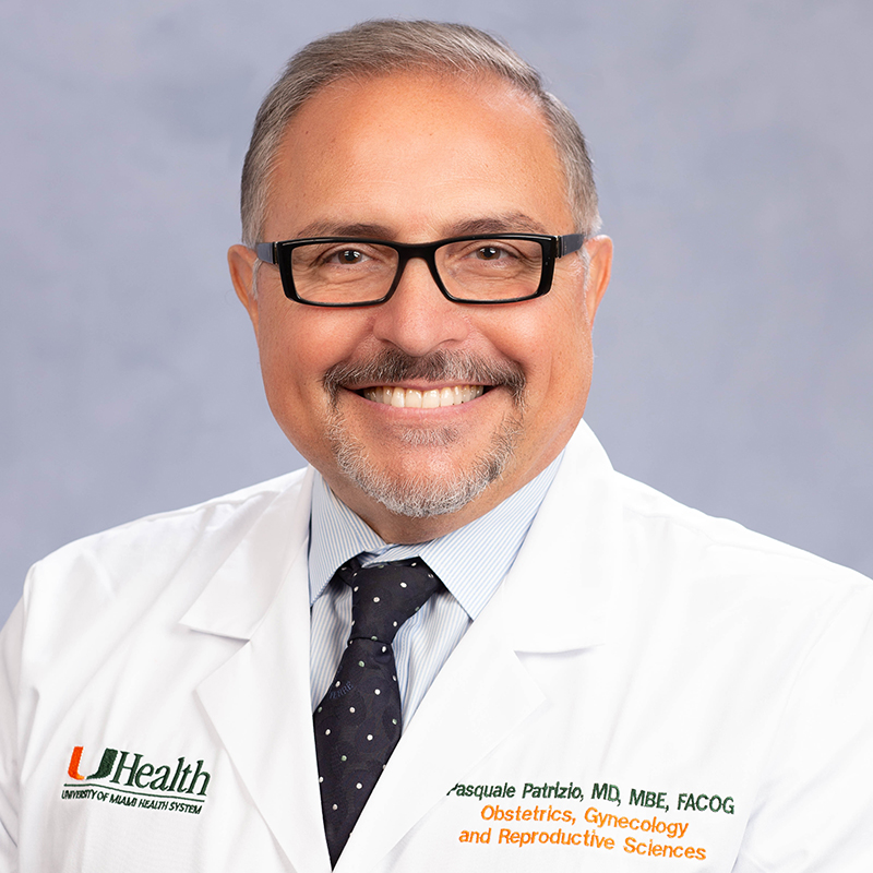 A headshot of a medical professional, he has on dark trimmed glasses, a white medial coat, blue collard dress shirt, and a dark blue tie