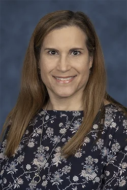A headshot of a woman with long brown hair, she smiles at the camera