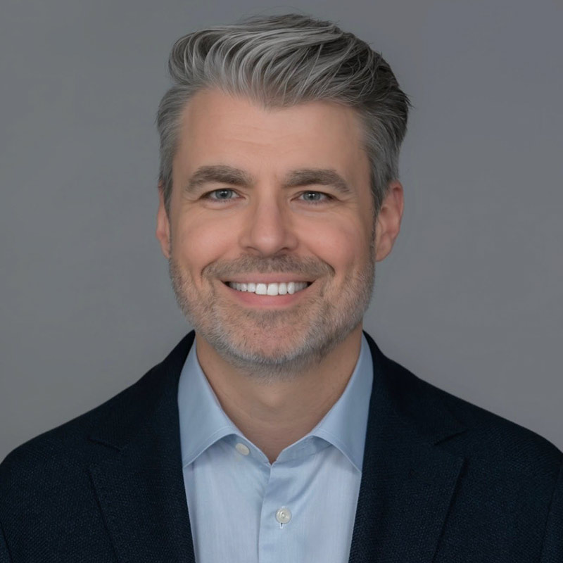 Closeup of a man, he smiles at the camera, has on a black coat and blue collar shirt