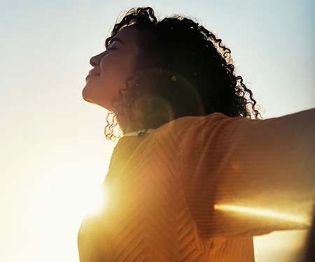 A woman outdoors, her eyes are closed, her arms are extended out, she is smiling