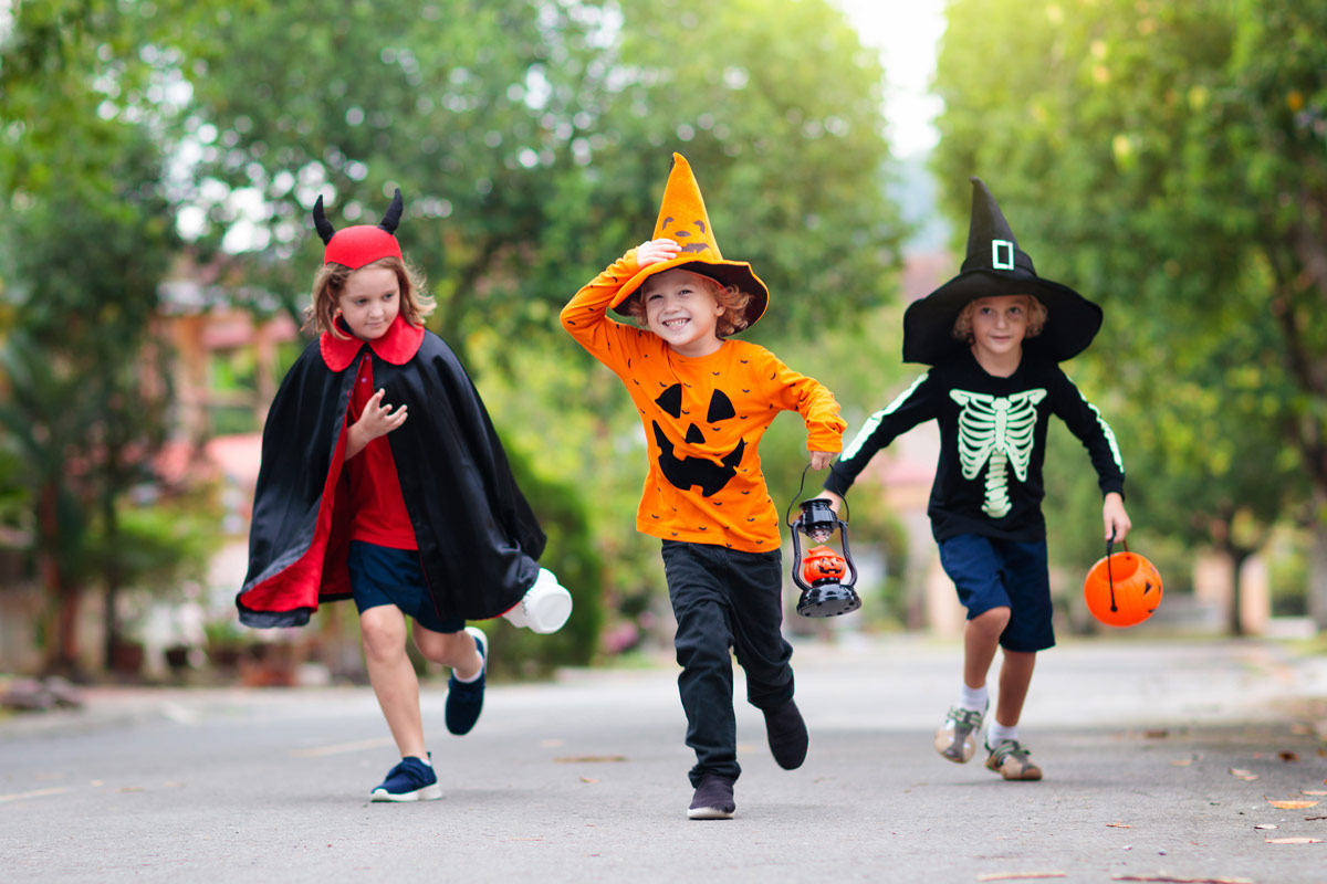 Three kids smiling and dressed up for Halloween walking on the street; one is dressed as a pumpkin, another a skeleton, and one a devil