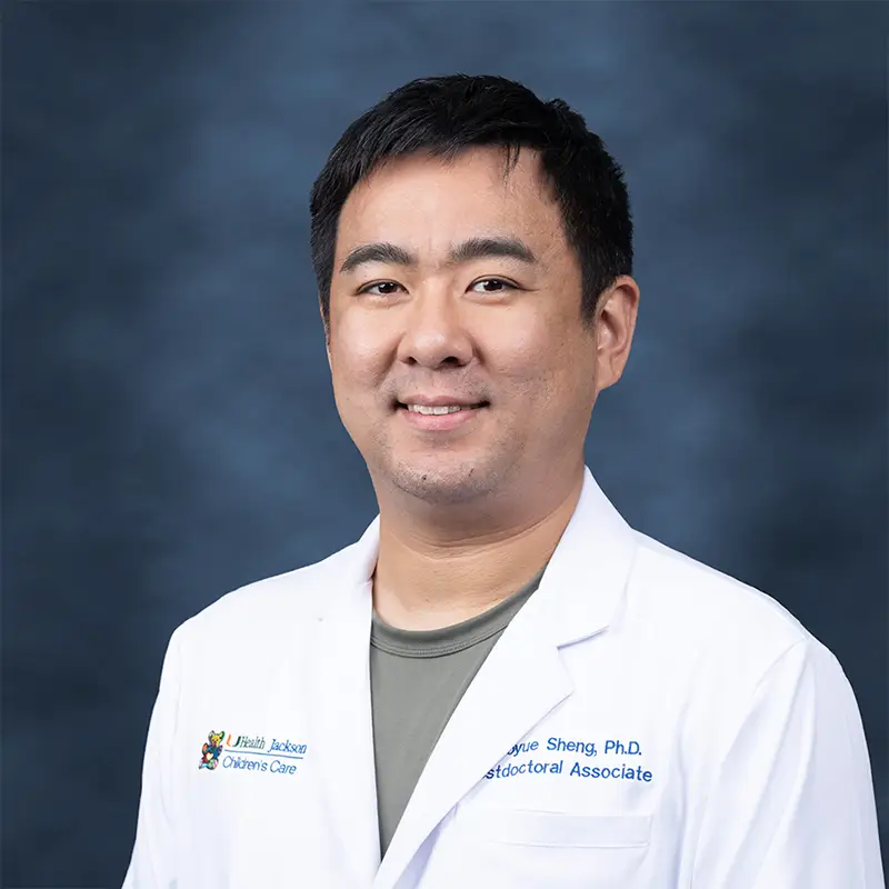 Professional headshot of Haoyue Sheng, Ph.D., Postdoctoral Associate, wearing a white lab coat with a UHealth Jackson Children's Care badge, smiling against a blue-gray studio background.