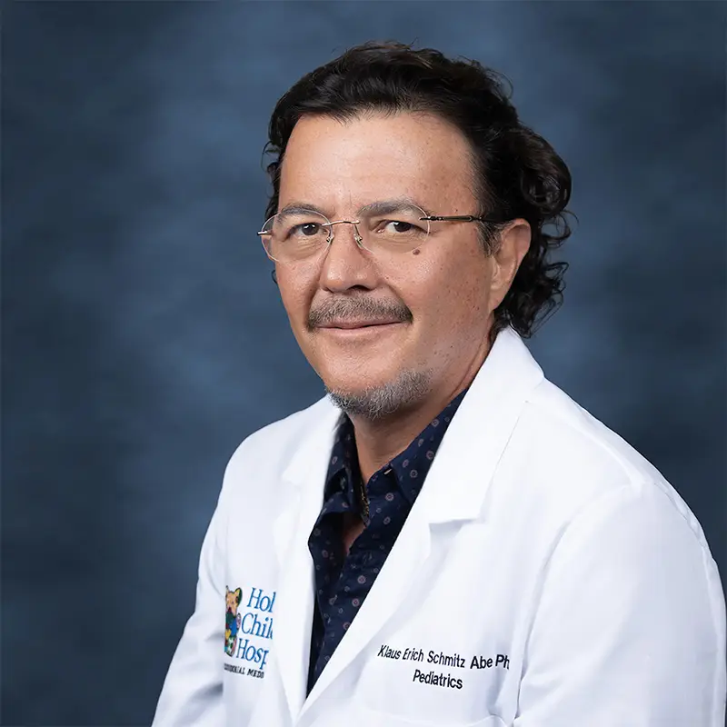 Professional headshot of Klaus Erich Schmitz-Abe, Ph.D., Pediatrics, smiling, wearing wire-rimmed glasses and a white lab coat with a Holtz Children's Hospital badge over a patterned shirt, against a blue-gray studio background
