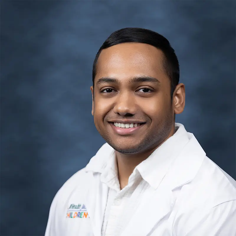Professional headshot of Madesh Ramesh smiling broadly, wearing a white lab coat with a UHealth Jackson Children's badge, against a blue-gray studio background