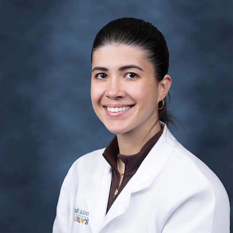 Professional headshot of Marel smiling, wearing a white lab coat with a UHealth Jackson Children's badge, hair pulled back, gold hoop earrings, against a blue-gray studio background