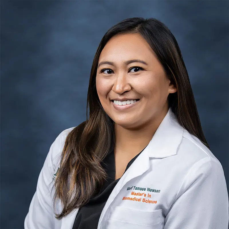 Professional headshot of Mari Tamase Newsam, Master's in Biomedical Sciences, smiling, wearing a white lab coat over a dark top, long dark hair, against a blue-gray studio background