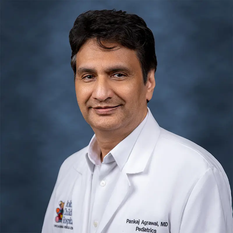 Professional headshot of Pankaj Agrawal, MD, Pediatrics, smiling, wearing a white lab coat with a Holtz Children's Hospital badge over a light collared shirt, against a blue-gray studio background