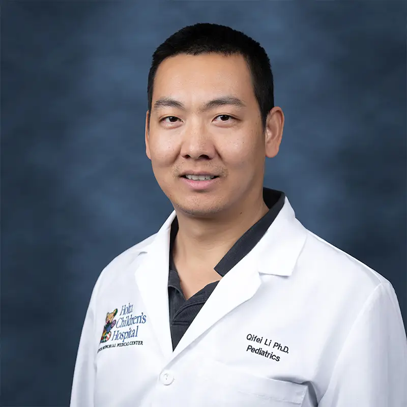 Professional headshot of Qifei Li, Ph.D., Pediatrics, smiling, wearing a white lab coat with a Holtz Children's Hospital badge over a dark polo shirt, against a blue-gray studio background