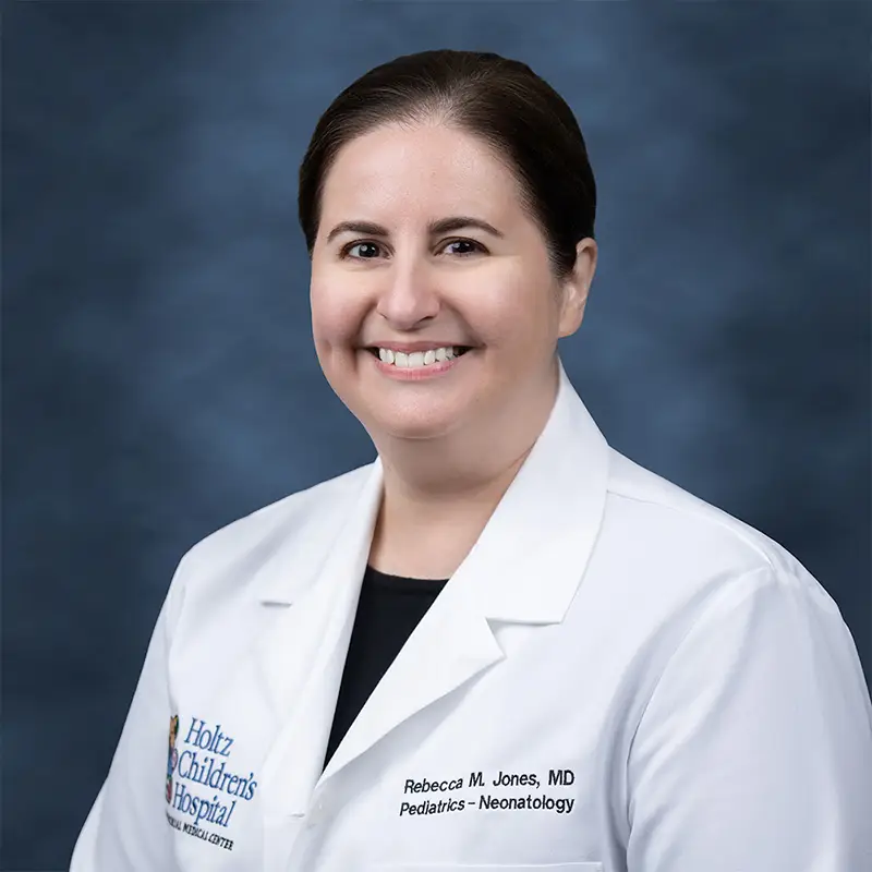 Professional headshot of Rebecca M. Jones, MD, Pediatrics–Neonatology, smiling broadly, wearing a white lab coat with a Holtz Children's Hospital badge over a dark top, hair pulled back, against a blue-gray studio background
