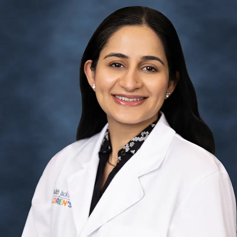 Professional headshot of Sara Ali smiling, wearing a white lab coat with a UHealth Jackson Children's badge over a patterned black top, against a blue-gray studio background