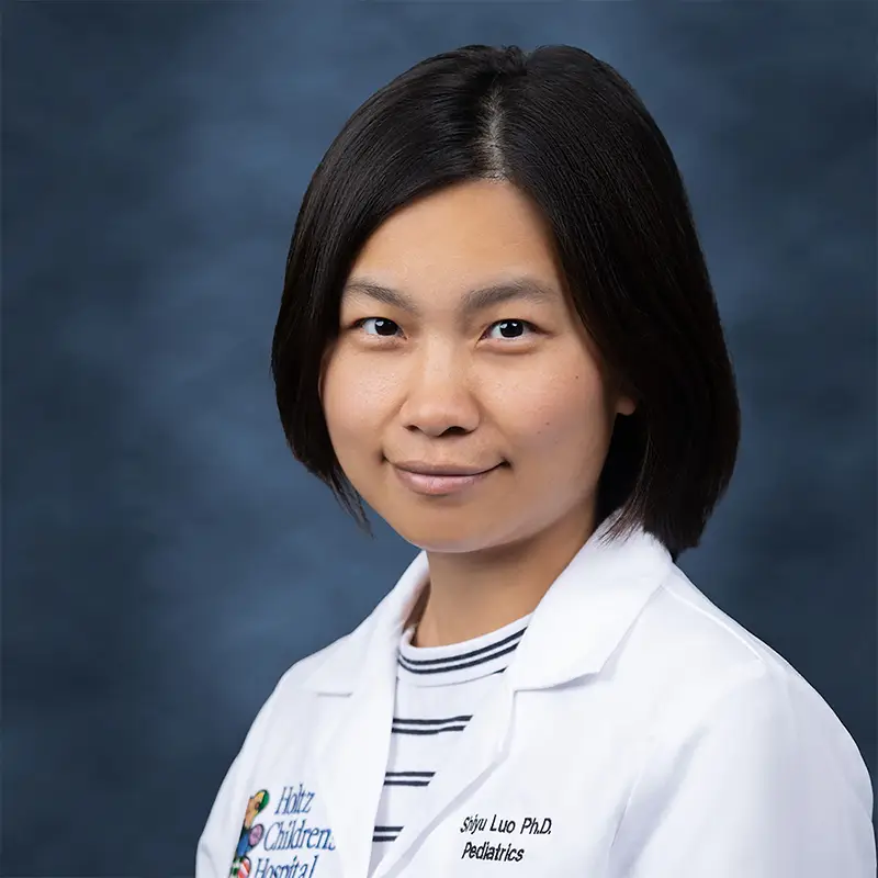 Professional headshot of Shiyu Luo, Ph.D., Pediatrics, smiling, wearing a white lab coat with a Holtz Children's Hospital badge over a striped turtleneck, against a blue-gray studio background