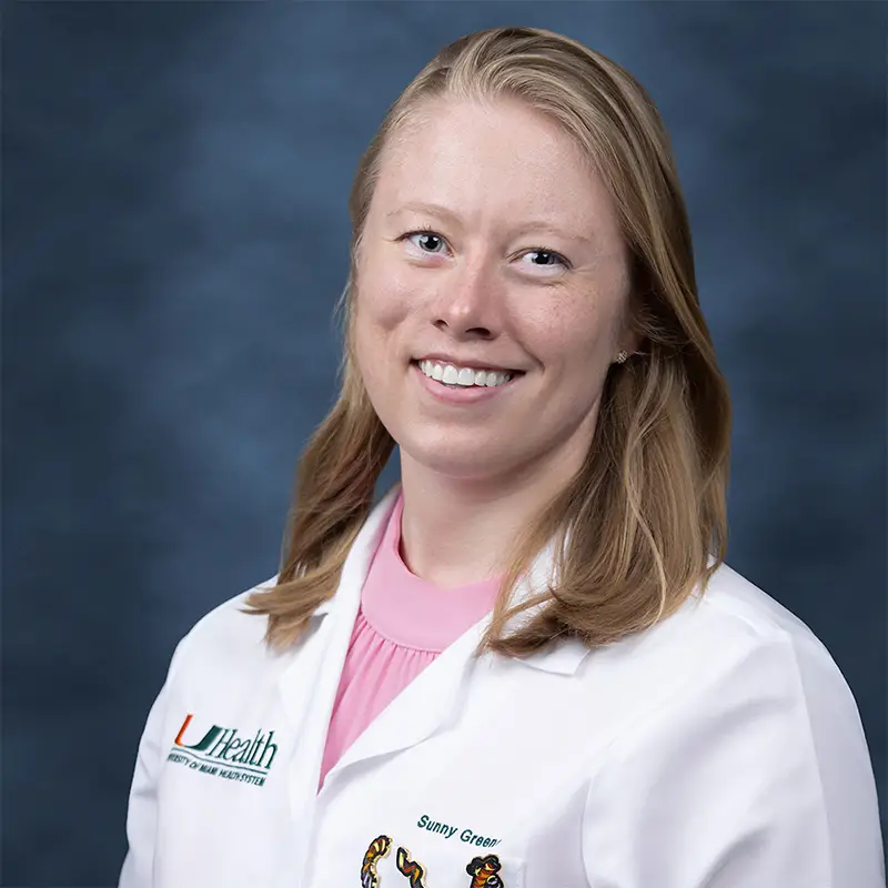 Professional headshot of Toni Marie Yeasky smiling, wearing a white lab coat with a UHealth Jackson Children's badge, hair pulled back, gold hoop earrings, against a blue-gray studio background