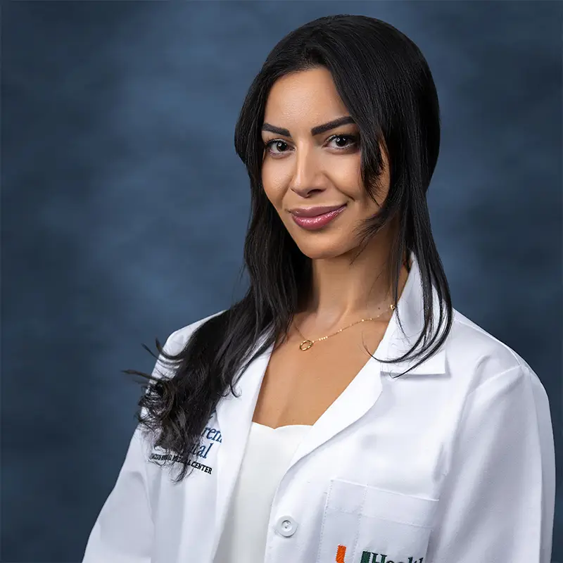 Professional headshot of Tzofia Drori smiling, wearing a white lab coat with a UHealth badge, long dark hair, gold necklace, against a blue-gray studio background