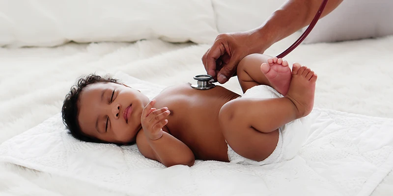 A baby lying on a white blanket is being examined with a stethoscope by a person's hand