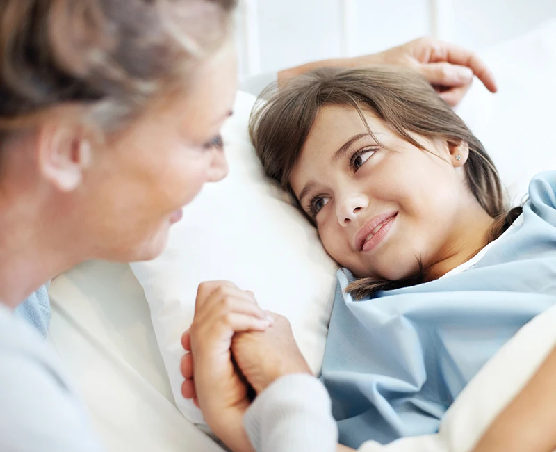 A closeup of a young girl laying on a hospital bed looking at an older woman and holding her hand