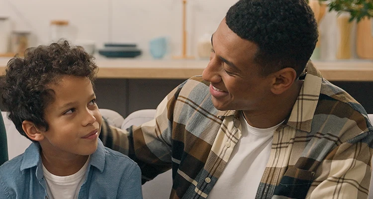 A dad speaking and smiling at his young son, they sit on a couch
