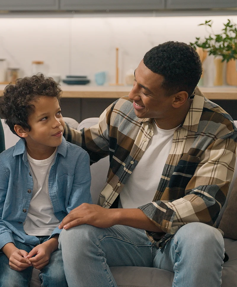 A dad speaking and smiling at his young son, they sit on a couch