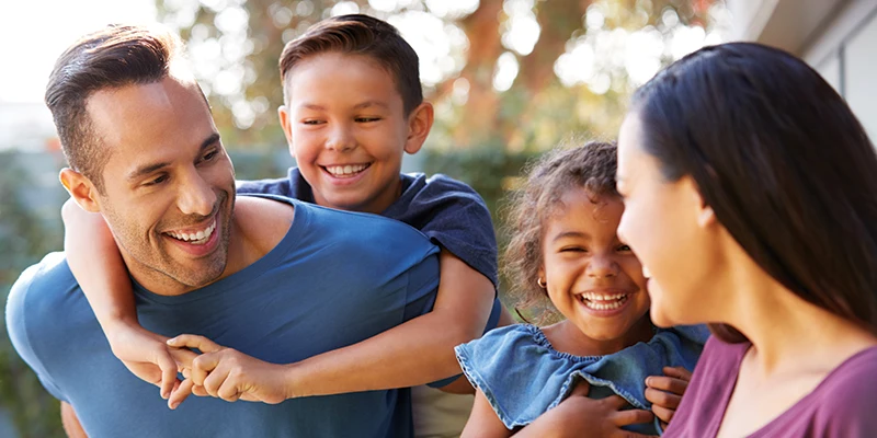 A happy family of four, including a father, mother, son, and daughter, smiling and interacting outdoors in a garden setting.