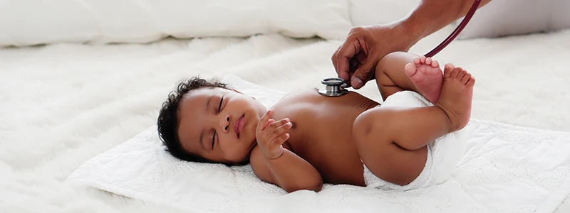 A baby lying on a white blanket is being examined with a stethoscope by a person's hand
