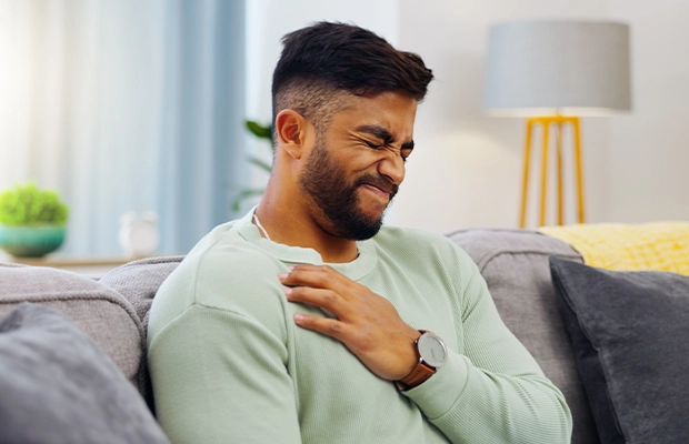 Man in a green suit sitting on a sofa holding his chest as if in severe pain