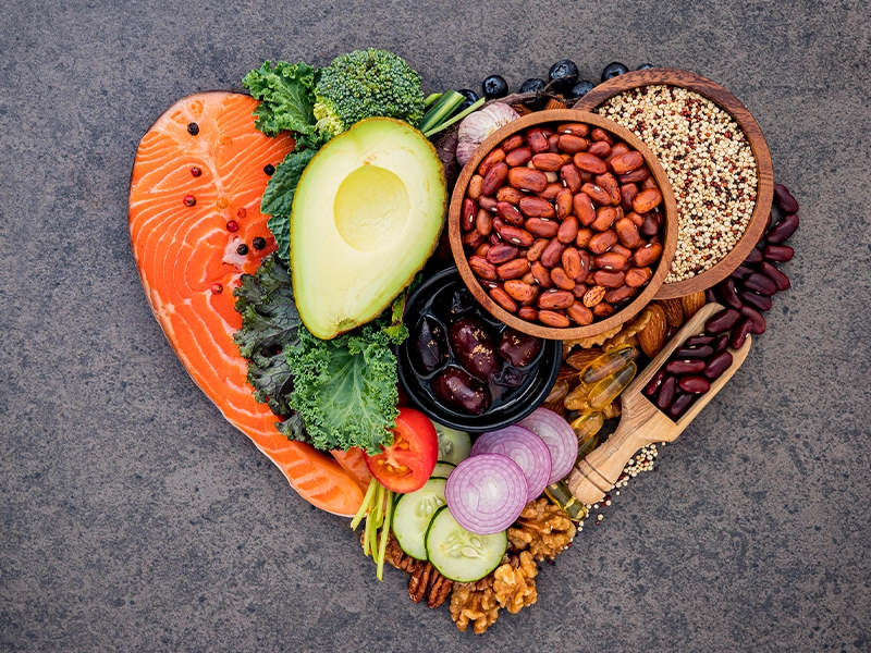 An image of food arranged in the shape of a heart; visible items include salmon, avocado, grains, onion, cucumber, and broccoli, among others.