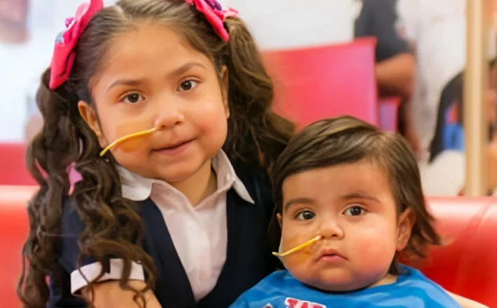 Two young children sitting together, they have a tube coming out of their noses