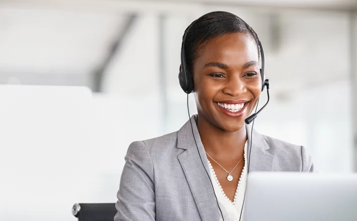 A woman smiling, she looks at her computer and has on a headset