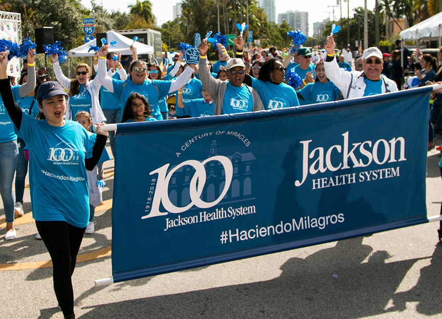 A group of people cheering, they are walking in a street, they all wear blue shirts and are holding a banner that reads A Century of Miracles Jackson Health System, Haciendo Milagros