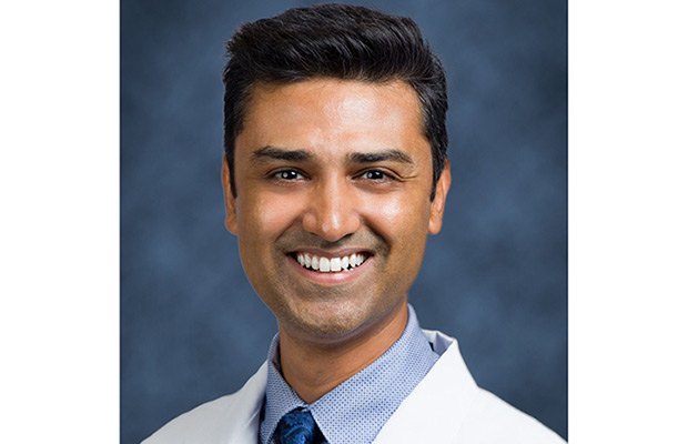 A man smiling at the camera, he is wearing a white coat, blue collard shirt, and a blue tie