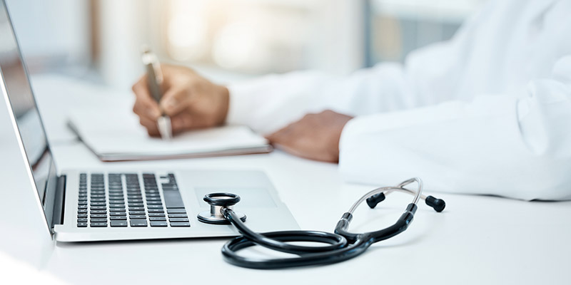 Closeup of a laptop, stethoscope, and doctor writing in a notebook