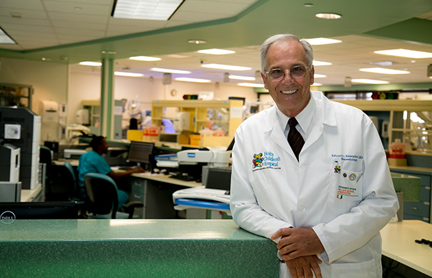 A physician smiling at the camera, he has on a white coat, a dark tie, and glasses