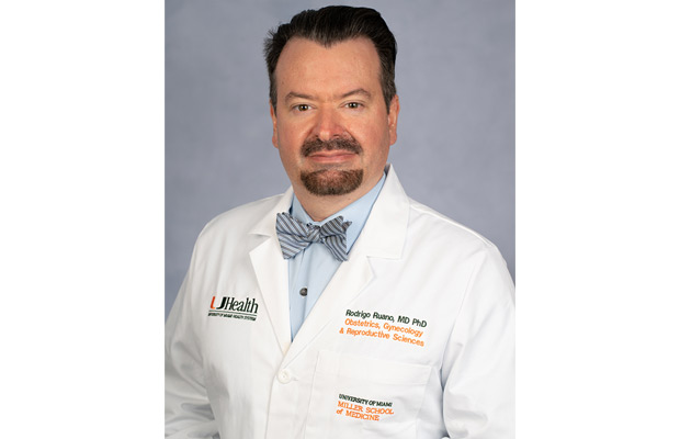 A headshot of a medical professional, he has on a white coat, light blue shirt, and bow tie, he looks at the camera