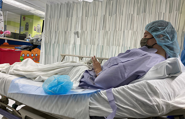 A young woman laying on a medical bed, she has on medical gown and cap