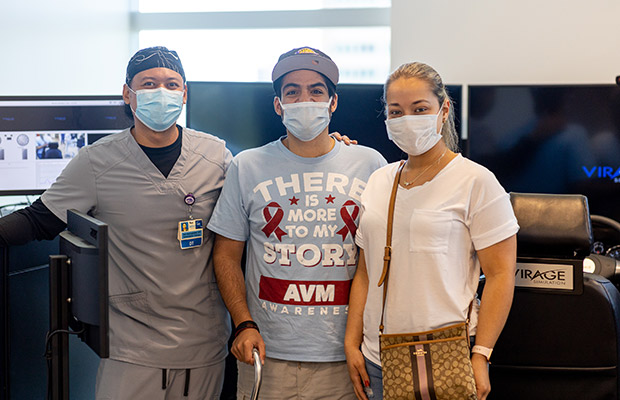 Three people standing in front of a driving simulator, they are all looking at the camera and wearing face masks