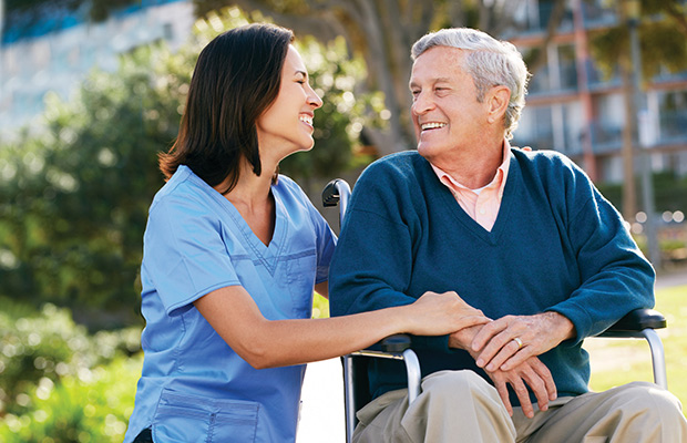 A nurse wearing blue scrubs is looking towards an older man who is sitting on a wheelchair, he looks back at her