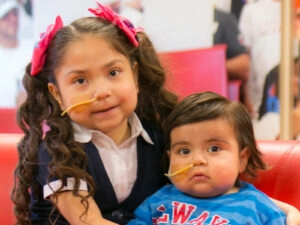 Two young children sitting next to each other, they have a tube coming out of their noses, they both look at the camera